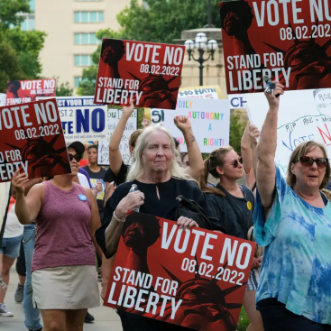 Activist march down a sidewalk holding signs that read "Vote No, Stand for Liberty"